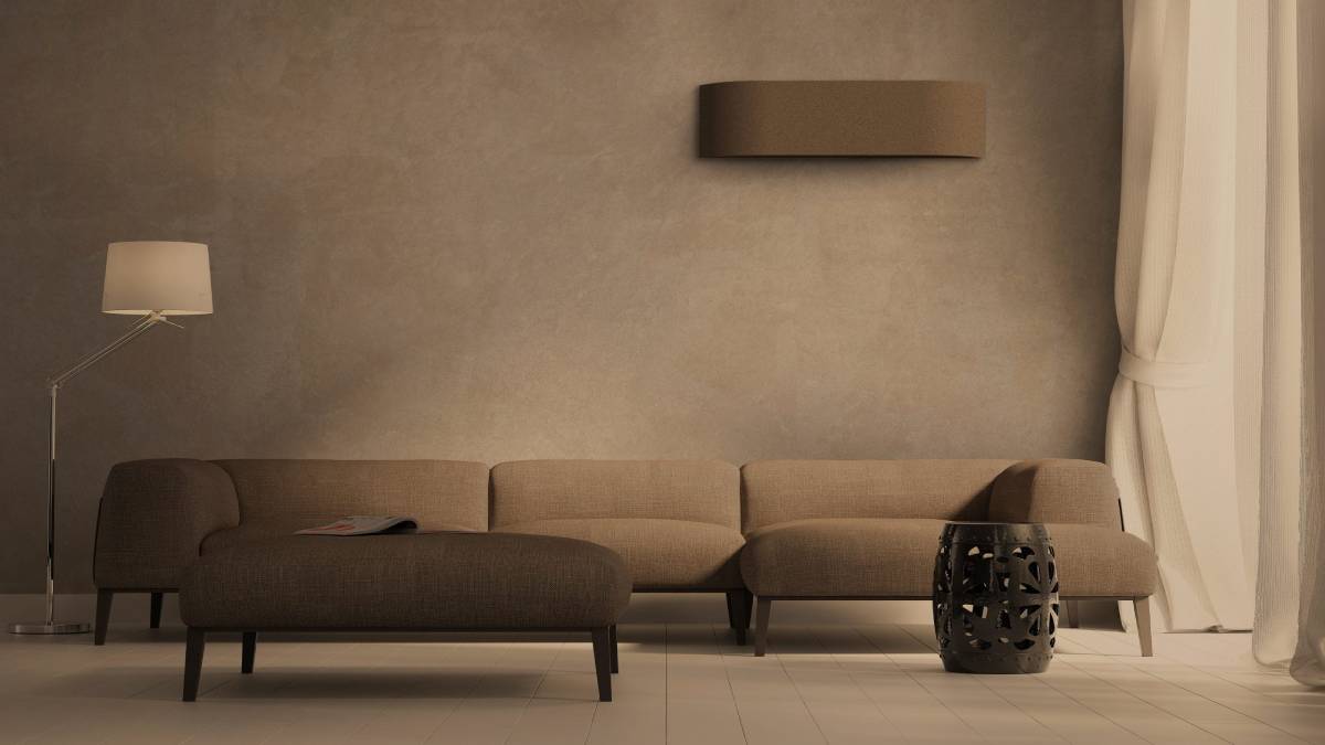 Een woonkamer met een bruine binnenunit van een design airco. In de ruimte staat een lange bruine zetel en een designlamp met witte lampenkap.
