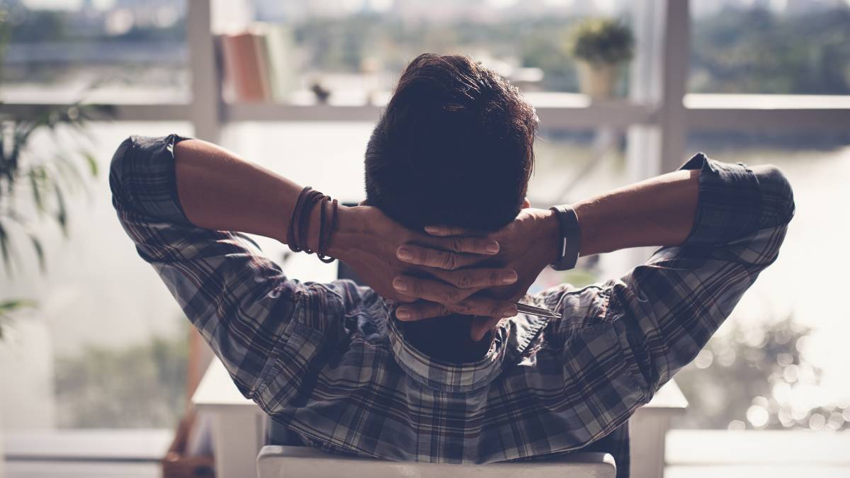 Een man in geruit hemd zit in een stoel en kijkt door een raam naar buiten. Hij heeft zijn handen achter zijn hoofd en leunt tevreden naar achter.