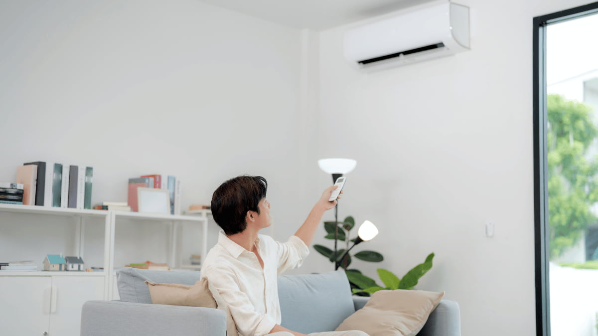 Een man zit op een lichtblauwe sofa. Hij bedient de binnenunit van zijn airco met een afstandsbediening.