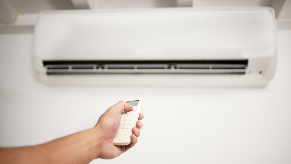 Een man zit op een grijze bank en bedient de witte binnenunit van de airco in huis met een afstandsbediening.