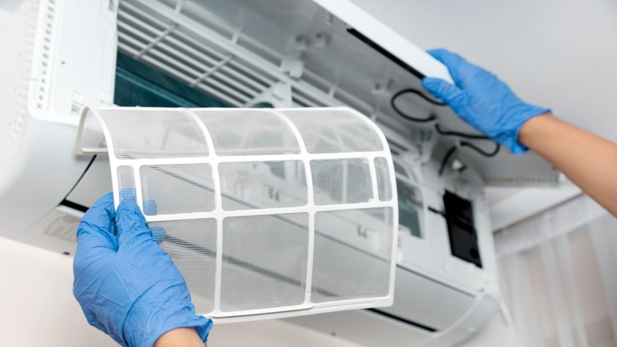 Een airco wordt geopend voor onderhoud. Twee handen met blauwe latex handschoenen halen de filter uit de airco.