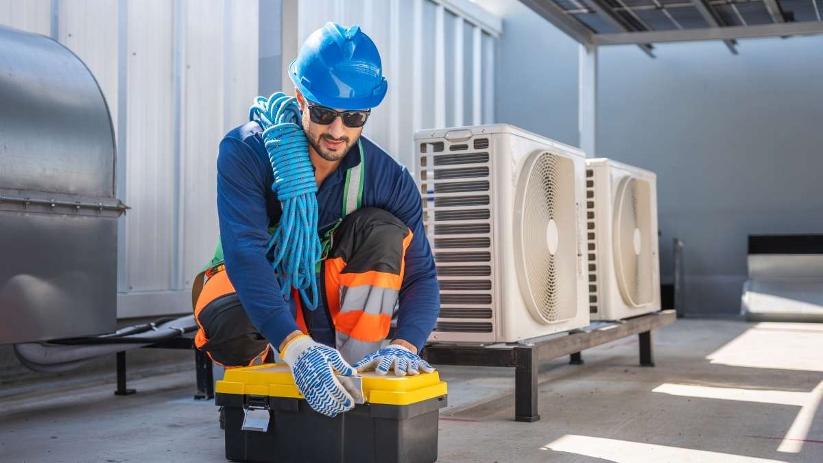 Een airco-installateur zit gehurkt op een dak met twee buitenunits. Hij heeft een gereedschapskist voor zich en een blauw touw om zijn schouder.