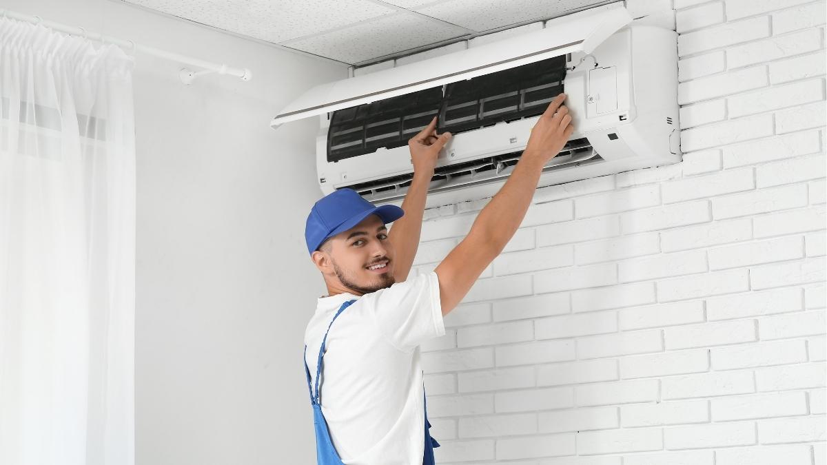 Een airco-installateur plaatst een witte Carrier binnenunit tegen een wit geschilderde bakstenen muur. Hij kijkt lachend over zijn schouder.