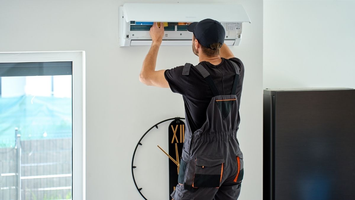 Een airco-installateur werkt aan de witte binnenunit van een aircosysteem. De binnenunit hangt in een witte woonkamer met raam, net boven een grote klok.