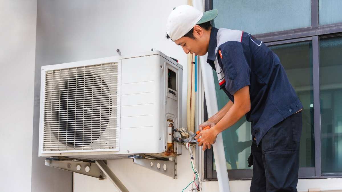 Een airco-installateur werkt aan de buitenunit van een airco. Hij draagt werkkledij en een pet.
