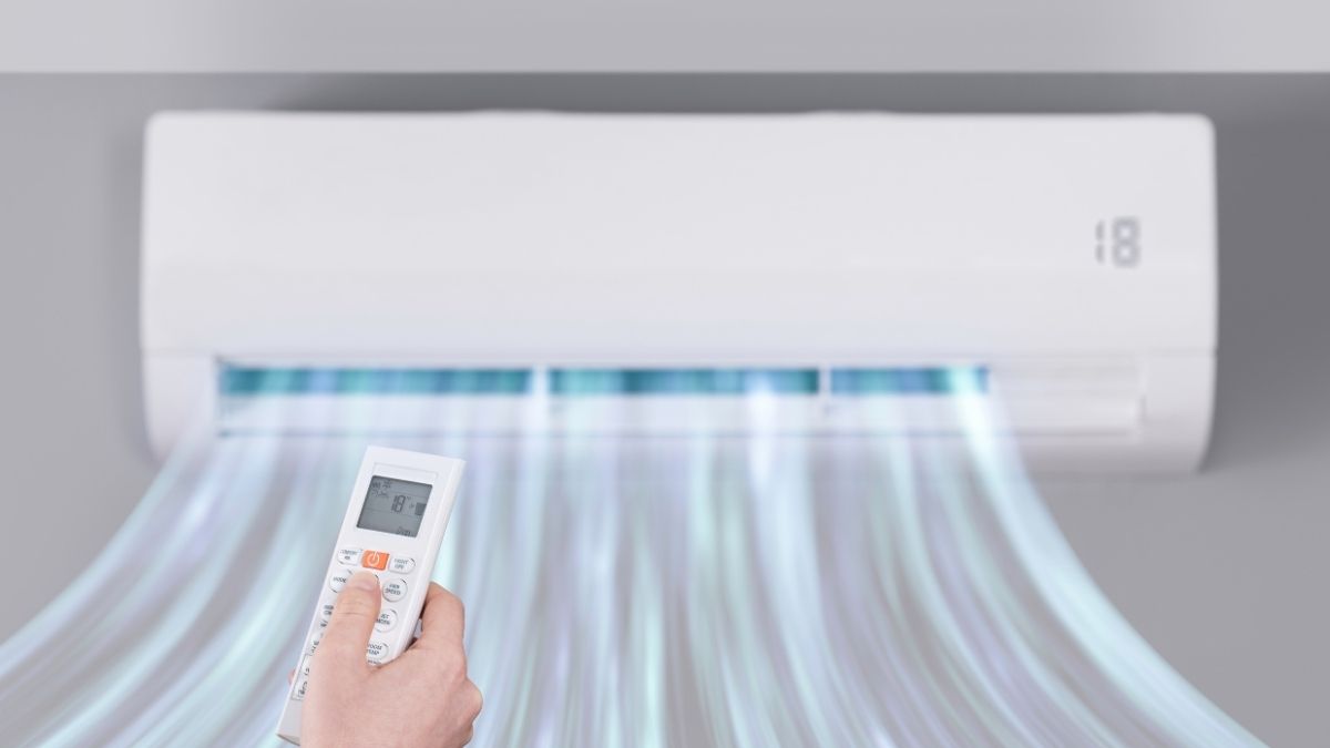 Een witte airconditioner verspreidt koele, blauwe lucht door het huis. Een hand met afstandsbediening stelt de gewenste temperatuur in.