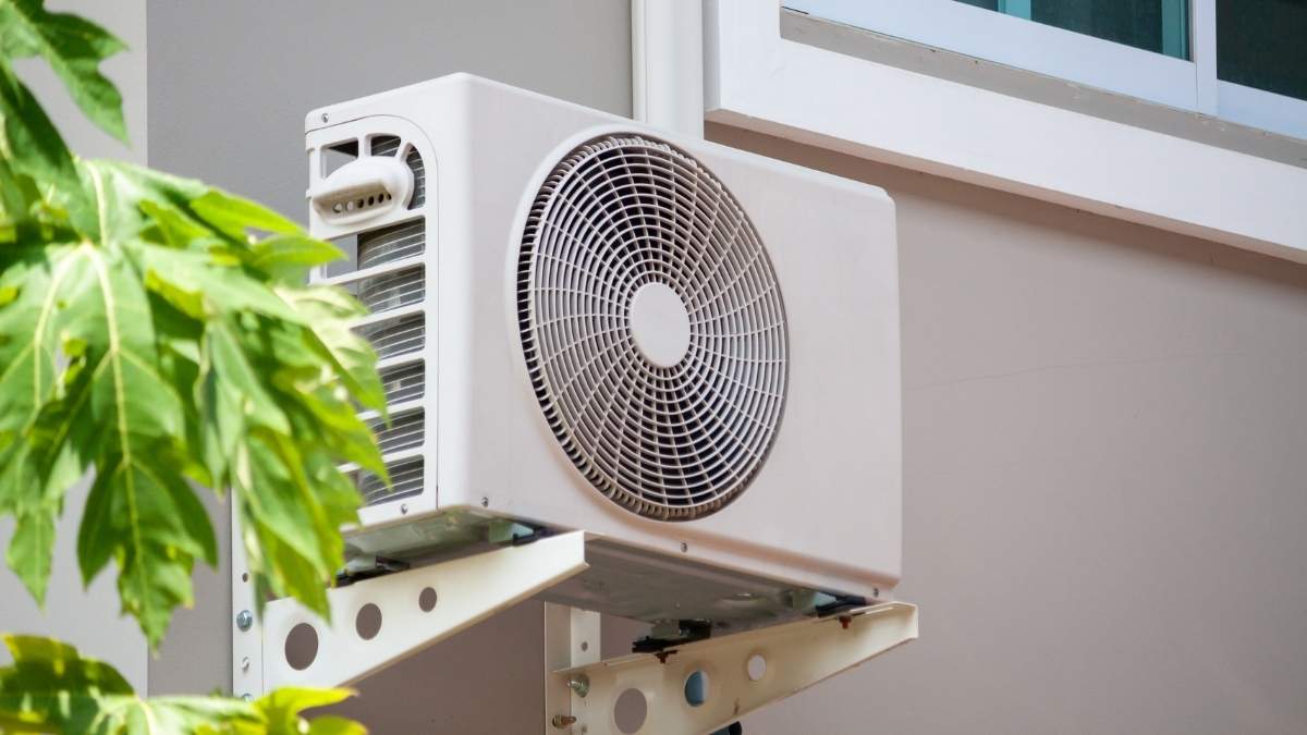Een buitenunit van een airco. De buitenunit heeft een witbeige kleur en is op stevige winkelhaken tegen een bruine gevel gemonteerd, net onder een raam.