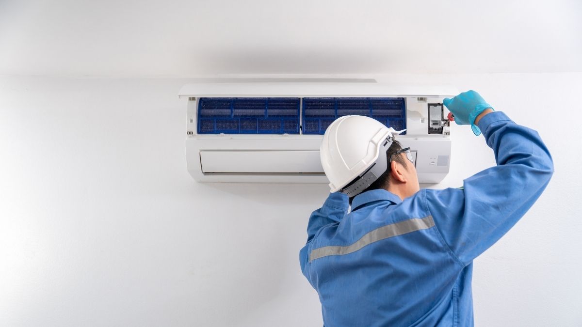 Een aircospecialist in blauwe werkkledij test een net geïnstalleerde witte airco binnenunit. De binnenunit hangt tegen een witte muur, net onder een wit plafond.