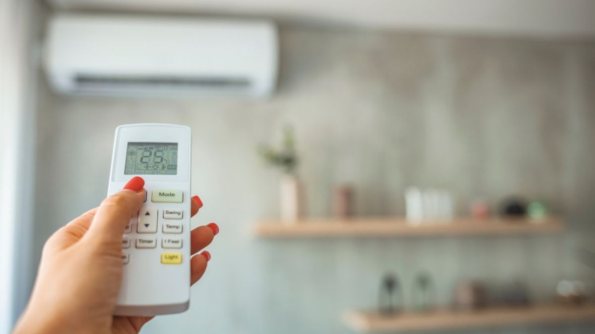 Een hand met roodgelakte nagels houdt een witte afstandsbediening vast. De afstandsbediening bestuurt een witte airco binnenunit die gemonteerd is tegen de wand van een keuken.