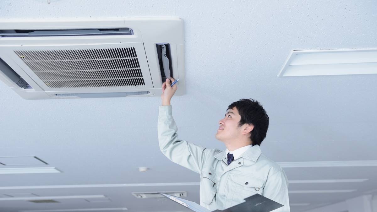 Een airco-installateur controleert het rooster van een ingebouwde cassette-airco. Hij draagt een witte jas en heeft een klembord vast.