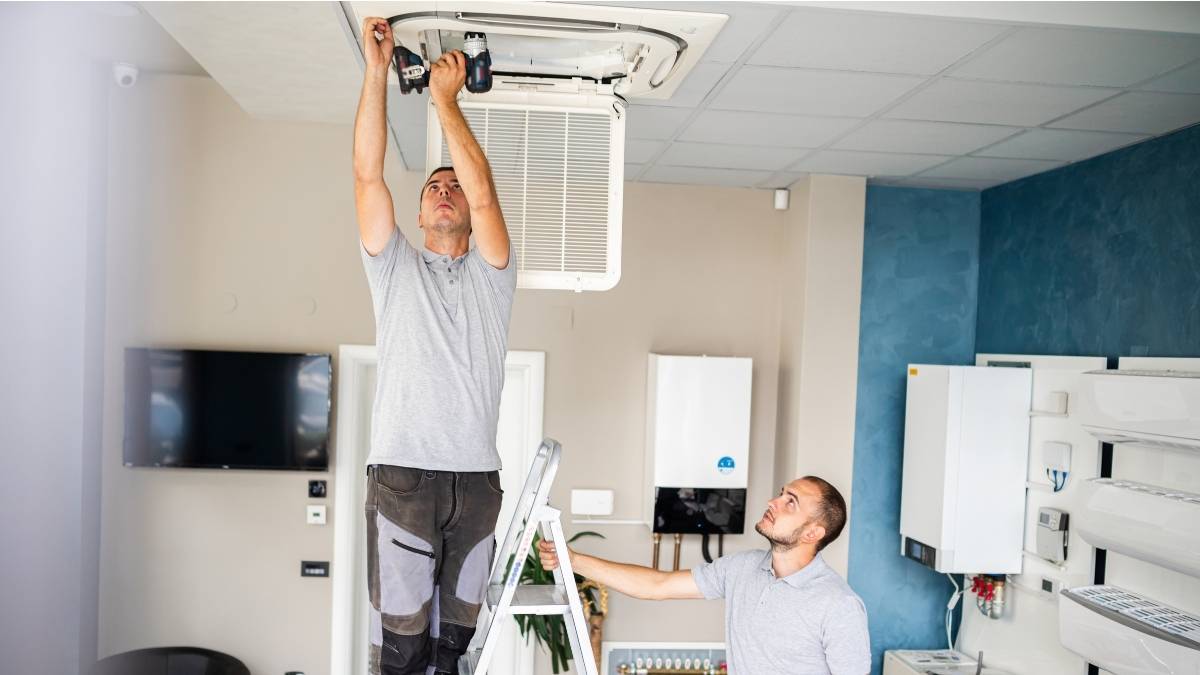 Twee airco-installateurs monteren een ingebouwde airco in het plafond van een woonkamer met open keuken. Een installateur staat op een ladder en gebruikt een boor. De andere houdt de ladder vast.