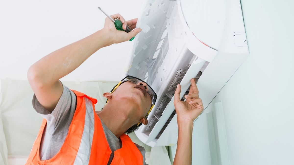 Een jonge airco-installateur in een oranje fluo-vest inspecteert een witte airco. Hij heeft een veiligheidsbril op en bekijkt de binnenunil langs onderen.