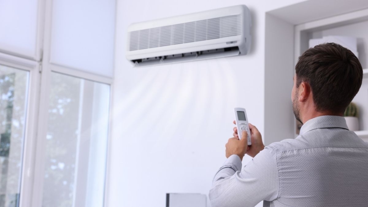 Een man in wit hemd regelt de temperatuur van zijn witte airco. De airco hangt tegen een witte muur, naast een hoog raam met zonwering.