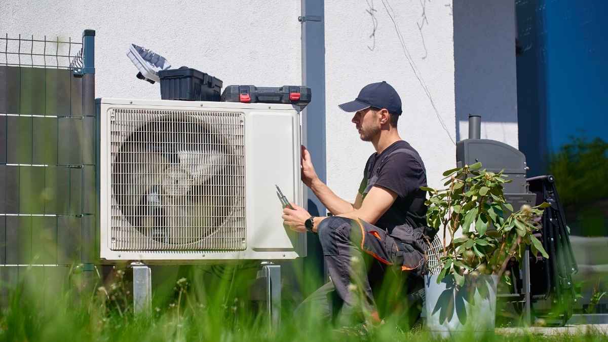Een airco-installateur plaatst de buitenunit van een airco tegen een witte gevel. Hij draagt werkkledij. Op de buitenunit liggen twee gereedschapskisten.