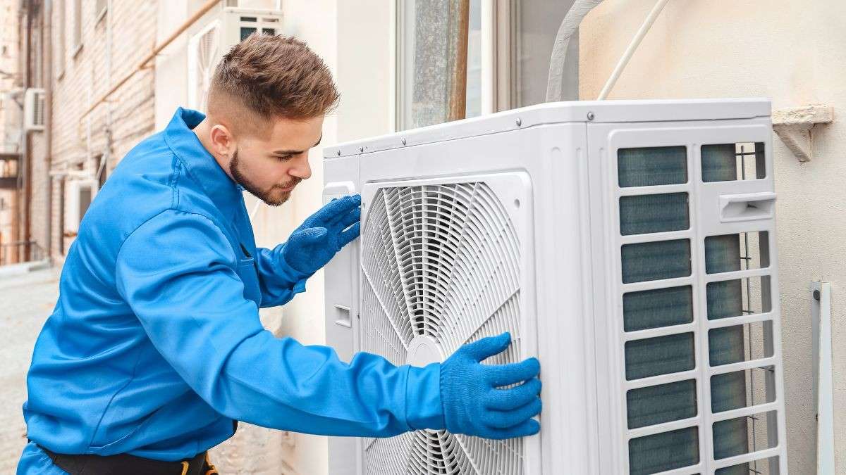 Een professionele installateur in blauwe werkkleding inspecteert de witte buitenunit van een airco die aan een gevel is gemonteerd.