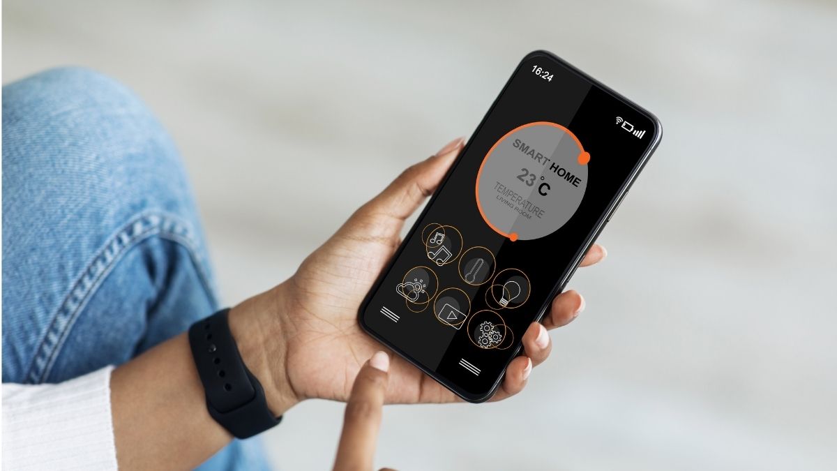 Een vrouw regelt de temperatuur in huis via een app op haar smartphone. Ze heeft een horloge, jeansbroek en witte trui aan.