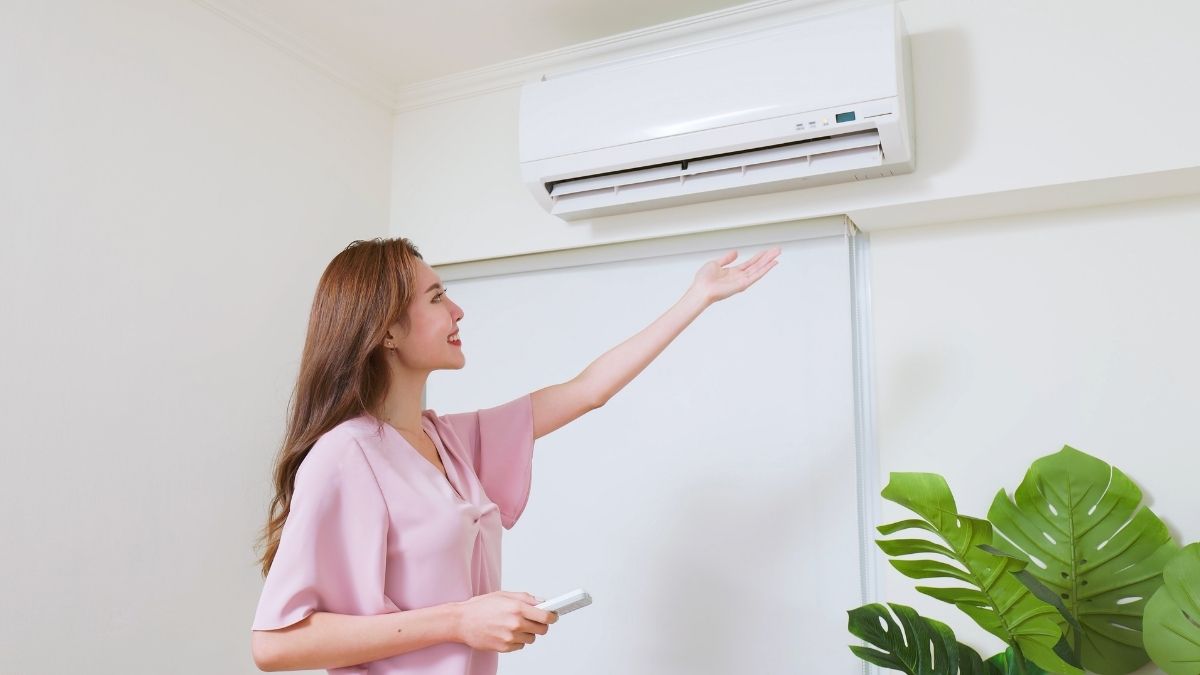 Een vrouw in een roze hemd steekt haar hand uit naar een airco binnenunit en voelt de koude lucht uit het wandmodel stromen. Onder de airco staat een kamerplant.