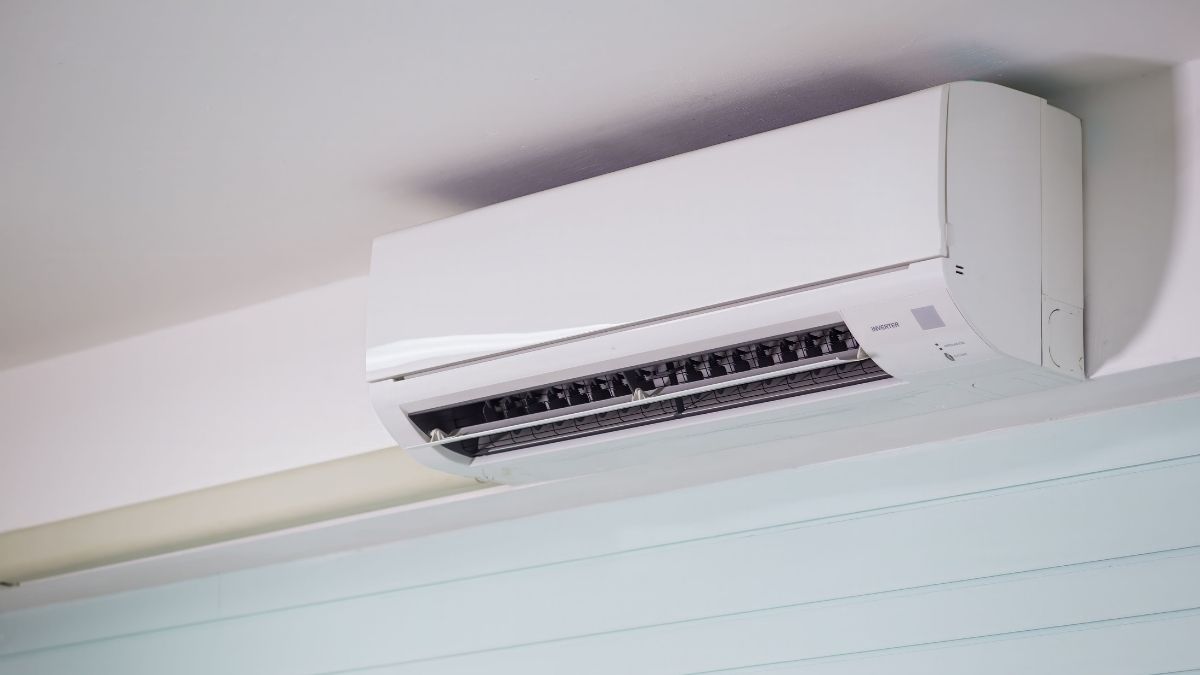 Een witte binnenunit in wandmodel, gemonteerd net onder het plafond, boven een raam met lichtblauwe lamellen.